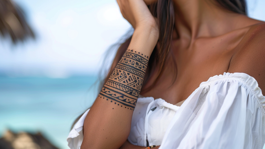 Femme portant une robe blanche avec un tatouage bracelet polynésien à motifs géométriques sur l'avant-bras, photographiée devant un océan turquoise